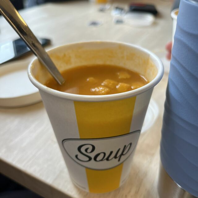 Suppe in einem Pappbecher, der weiß und gelb gestreift ist und auf dem "Soup" steht. Die Suppe ist rot und hat Croutons drin.