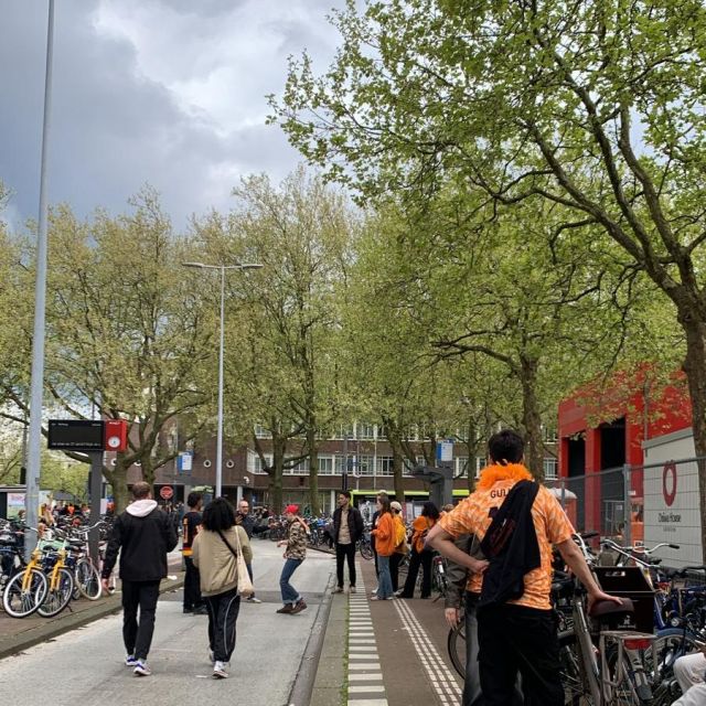Blick auf eine Straße. Links und rechts stehen Fahrräder. Im Hintergrund sind grüne Bäume. Man sieht einige Personen in orange gekleidet.
