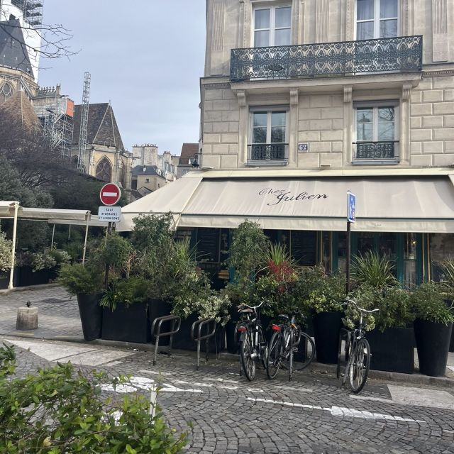 Blick auf eine Straße in Paris. Auf der gegenüberliegenden Seite ist ein Restaurant, dass von einer Markise überspannt wird. Es stehen viele Büsche in Kübeln um das Restaurant herum.