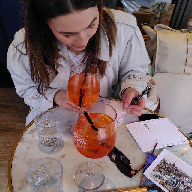 Eine Person sieht an einem Tisch mit zwei orangefarbenen Getränken. Sie blickt auf den Tisch und hält einen Stift in der linken Hand. Vor ihr liegen mehrere Postkarten.