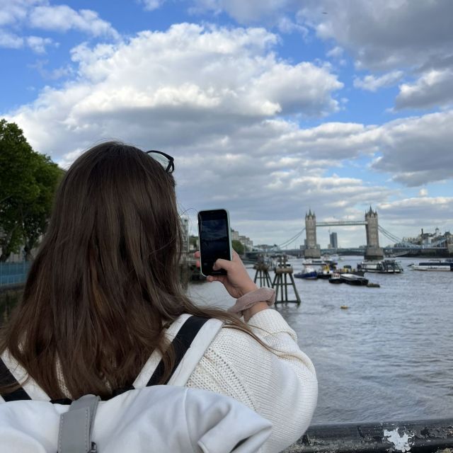 Blick auf eine Person mit Rucksack von hinten, die mit dem Smartphone etwas fotografiert. Im Hintergrund sieht man einen Fluss und in der Ferne eine Brücke mit zwei imposanten Pfeilern (die Tower Bridge).