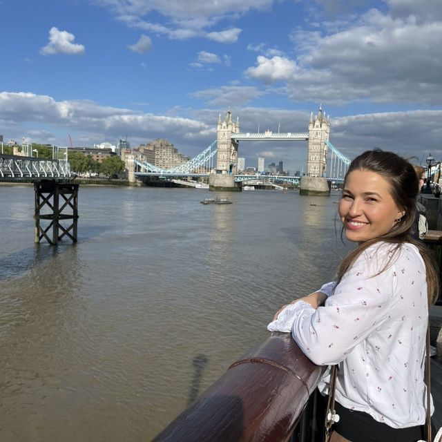 Eine Person lächelt in die Kamera. Sie steht an einem Geländer an einem Fluss, der Kopf zur Kamera geneigt. Sie trägt eine weiße Bluse. Im Hintergrund sieht man die Tower Bridge.