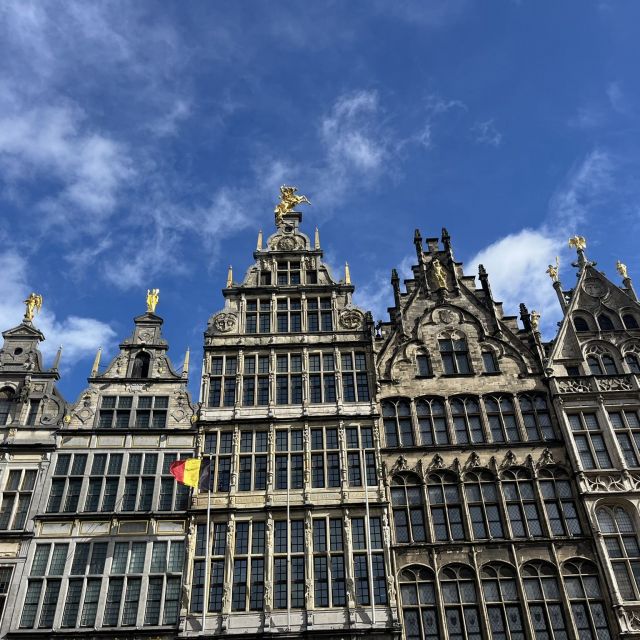 Der Himmel ist blau mit ein paar Wolken. Im Vordergrund steht ein altes mittelalterliches Gebäude, vor dem die belgische Fahne weht.
